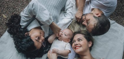 Family of four enjoying time together on a blanket outside, smiling and relaxed.
